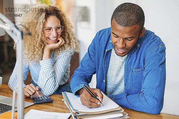 Junger Mann schreibt auf Papier  während er mit glücklicher Frau im Heimbüro sitzt