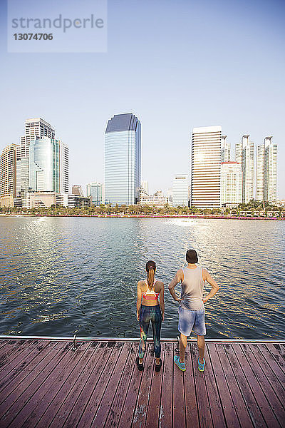 Rückansicht eines Paares  das auf einem Holzsteg am Fluss vor dem Stadtbild steht
