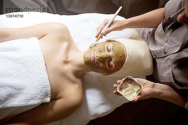 Draufsicht einer Therapeutin  die im Spa eine Gesichtsmaske auf das Gesicht einer Frau aufträgt