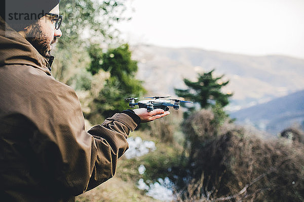 Seitenansicht eines männlichen Wanderers  der einen Quadkopter hält  während er auf einem Berg im Wald steht