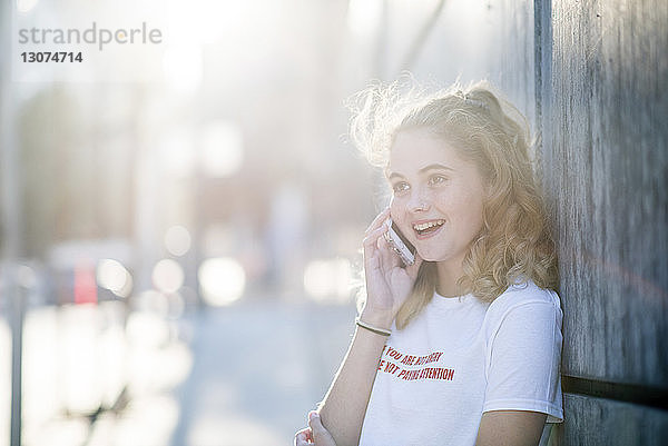 Fröhliche Teenagerin schaut weg  während sie mit einem Smartphone gegen eine Wand in der Stadt telefoniert