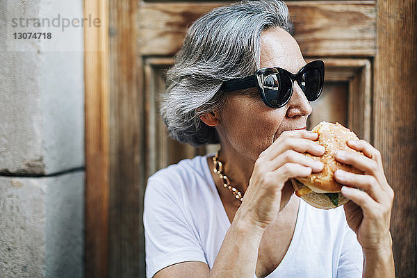 Frau mit Sonnenbrille isst Burger  während sie an der Tür sitzt