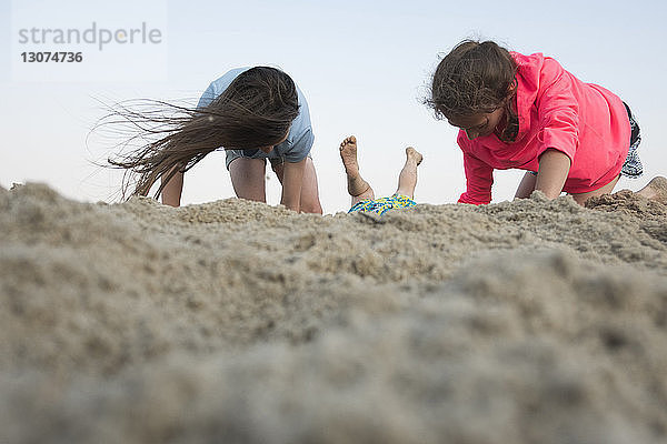 Ebenerdiges Bild von Schwestern  die mit Sand am Strand vor klarem Himmel spielen