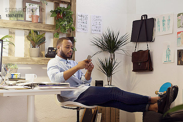 Kreativer Geschäftsmann in voller Länge  der ein Smartphone benutzt  während er im Büro mit erhobenen Füßen sitzt