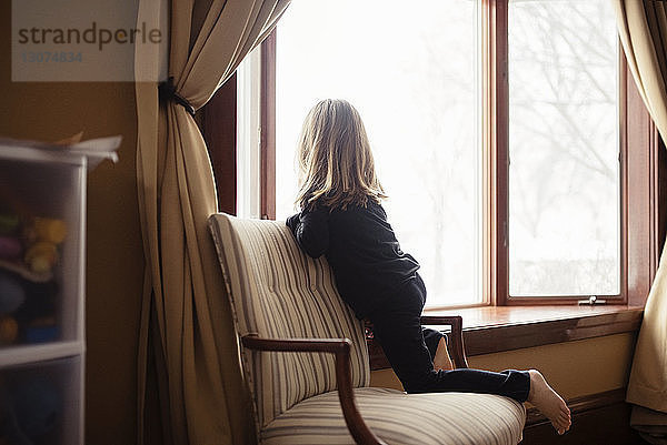 Rückansicht eines zu Hause auf einem Sessel knienden Mädchens am Fenster