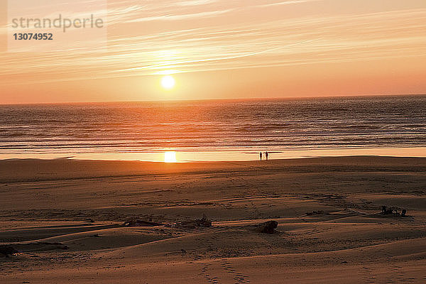 Entfernte Silhouette von Freundinnen  die bei Sonnenuntergang am Strand gegen den Himmel stehen