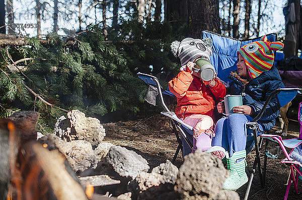 Fröhliche Schwestern trinken  während sie auf einem Campingplatz im Wald sitzen