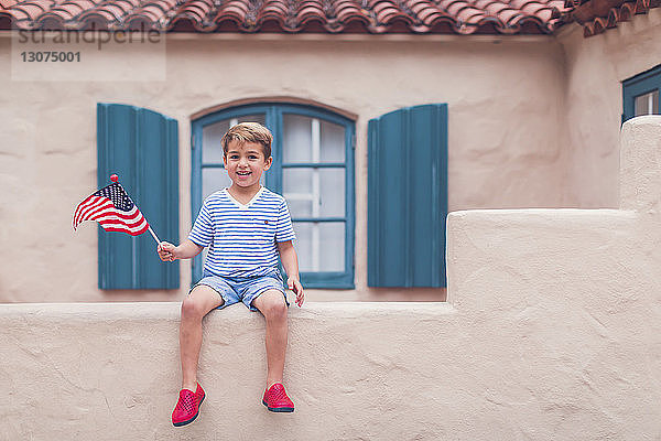 Porträt eines glücklichen Jungen  der die amerikanische Flagge hält  während er auf einer Stützmauer gegen das Haus sitzt