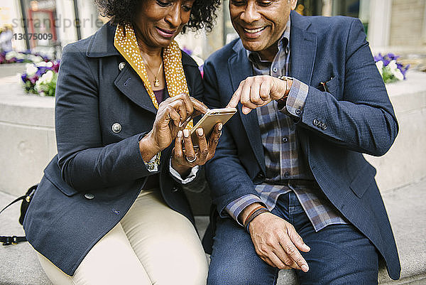Lächelndes Paar benutzt Smartphone  während es in der Stadt auf einem Sitzplatz sitzt