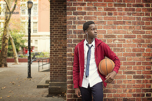 Nachdenklicher Student mit Basketball  der Musik hört  während er am Basketballplatz an einer Ziegelmauer steht