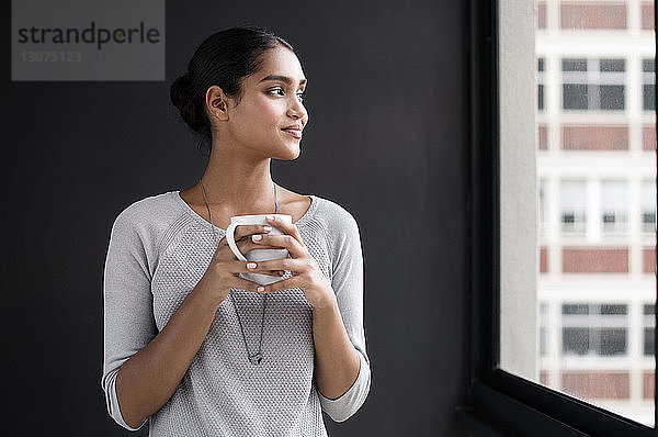 Lächelnde Innenarchitektin mit Kaffeebecher in der Hand  während sie im Büro durchs Fenster schaut