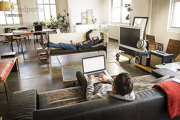 Hochwinkelansicht eines Geschäftsmannes mit Laptop  während ein Mitarbeiter im Kreativbüro auf dem Sofa liegt