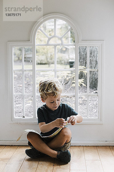 Junge mit Buch sitzt zu Hause auf dem Boden gegen das Fenster