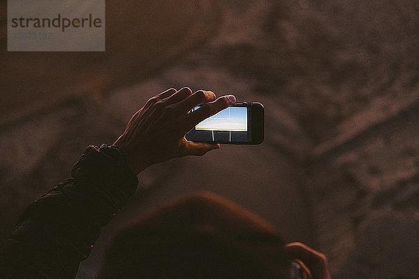 Abgetrennte Hand eines Mannes  der mit seinem Handy am Strand bei Sonnenuntergang fotografiert