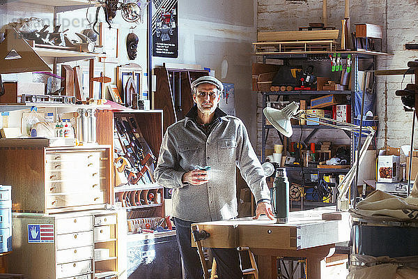 Porträt eines selbstbewussten Tischlers  der Getränke hält  während er in der Werkstatt am Tisch steht