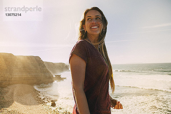 Lächelnde Frau schaut weg  während sie am Strand gegen den Himmel steht