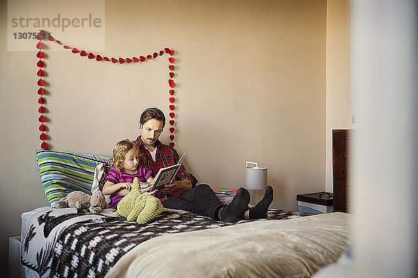 Vater zeigt einem Mädchen ein Buch  während er zu Hause auf dem Bett sitzt