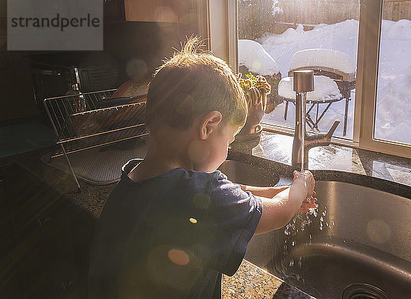 Junge wäscht sich im Winter die Hände am Küchenspülbecken am Fenster