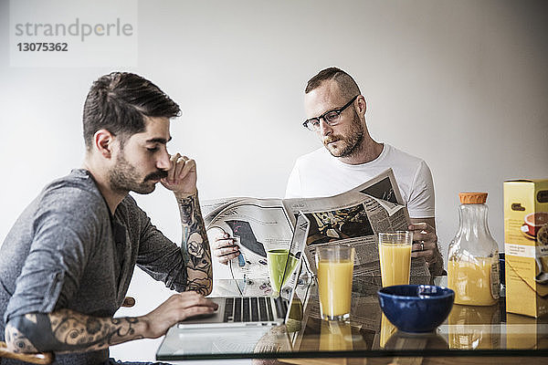 Schwuler Mann benutzt Laptop  während der Partner am Frühstückstisch Zeitung liest