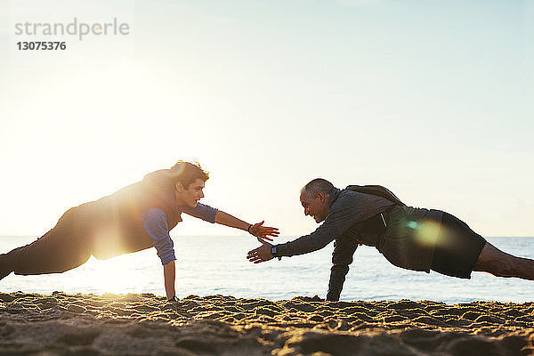 Seitenansicht von Vater und Sohn beim gemeinsamen Liegestützen gegen den klaren Himmel am Strand bei Sonnenuntergang