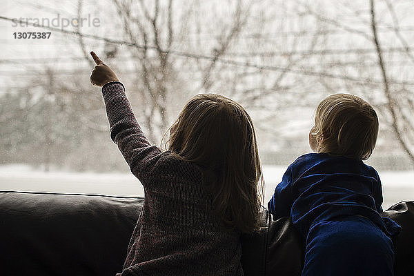 Rückansicht von Geschwistern  die durch das Fenster schauen  während sie sich im Winter zu Hause auf dem Sofa entspannen