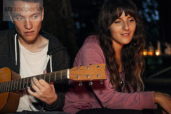 Mann spielt Gitarre  während er nachts mit seiner Freundin im Wald sitzt
