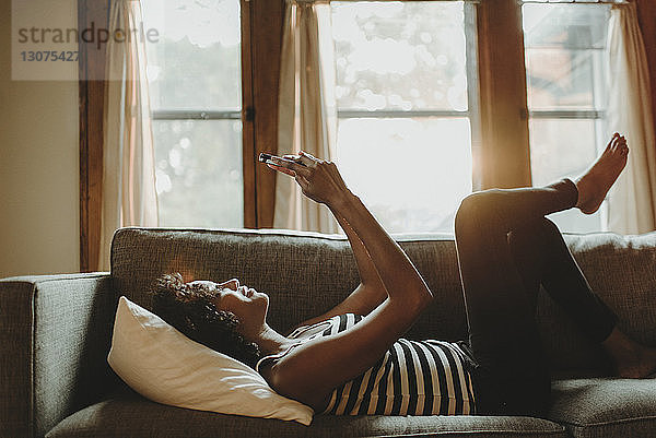 Seitenansicht einer Frau  die ein Smartphone benutzt  während sie zu Hause auf dem Sofa vor dem Fenster liegt