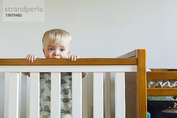 Süßer Babyjunge beißt auf Holzgeländer  während er zu Hause in der Krippe steht