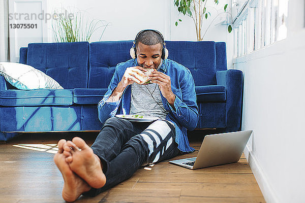 Mann isst Sandwich  während er zu Hause auf dem Sofa am Laptop Musik hört