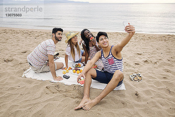 Mann nimmt Selfie mit Freunden mit  während er ein Picknick am Strand genießt