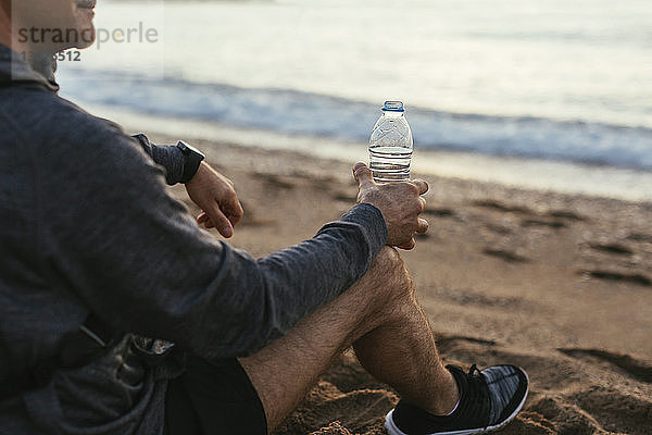 Niedriger Teil eines Mannes  der eine Wasserflasche hält  während er am Strand sitzt