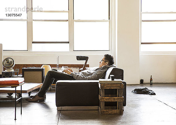 Seitenansicht eines Geschäftsmannes  der einen Laptop benutzt  während er im Büro auf dem Sofa liegt