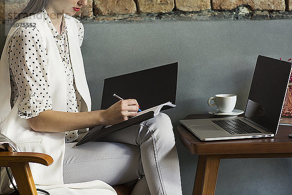 Mittelsektion einer Geschäftsfrau  die mit einem Laptop an einem Tisch in der Hotellobby in eine Akte schreibt