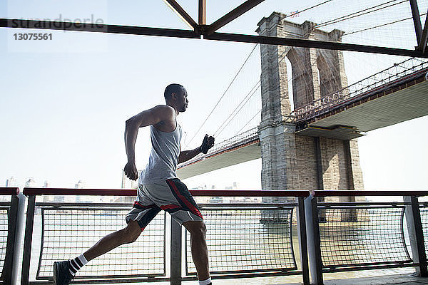 Seitenansicht eines jungen Mannes  der am Geländer gegen die Brooklyn Bridge joggt