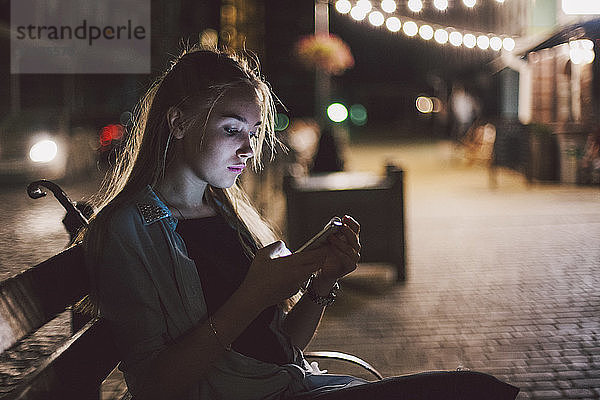 Junge Frau benutzt ein Smartphone  während sie nachts auf einer Bank am beleuchteten Bürgersteig in der Stadt sitzt