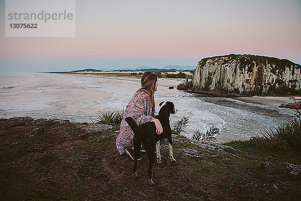 Seitenansicht einer Frau  die bei Sonnenuntergang mit einem Hund auf einem Hügel am Strand kauert