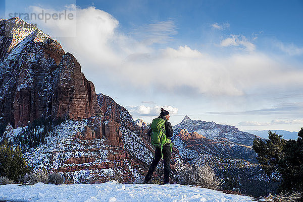 Rückansicht eines Mannes mit grünem Rucksack  der auf Schnee in der Wüste vor Felsformationen und bewölktem Himmel im Winter steht