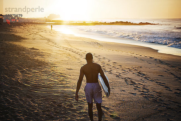 Mann trägt ein Surfbrett  während er am Strand auf Sand läuft