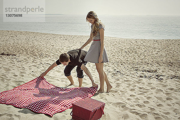 Freundin schaut Mann an  der am Strand eine Picknickdecke auf den Sand legt