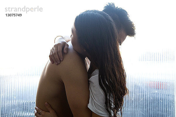 Seitenansicht eines romantischen Paares  das sich umarmt  während es zu Hause am Fenster steht