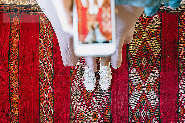Niedriger Teil einer Frau  die mit einem Smartphone fotografiert  während sie auf einem traditionellen Teppich steht