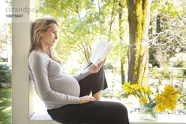 Schwangere Frau liest ein Buch  während sie sich an einem sonnigen Tag auf der Veranda ausruht