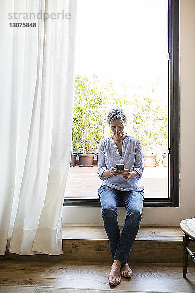Reife Frau in voller Länge  die ein Smartphone benutzt  während sie zu Hause auf dem Fensterbrett sitzt