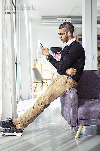 Seitenansicht eines Geschäftsmannes  der ein Smartphone benutzt  während er sich im Büro auf ein Sofa lehnt