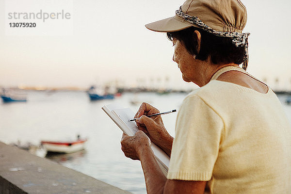 Ältere Frau zeichnet auf Buch  während sie an einer Stützmauer am Meer sitzt