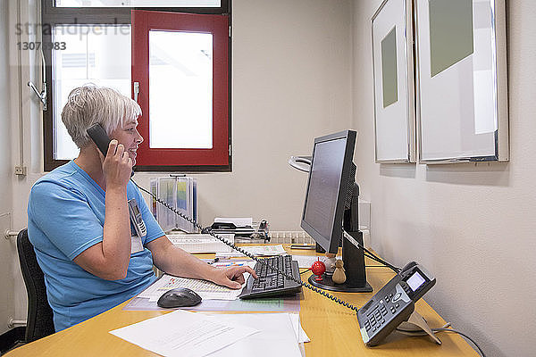 Seitenansicht einer Ärztin  die telefoniert  während sie einen Desktop-Computer im Krankenhaus benutzt