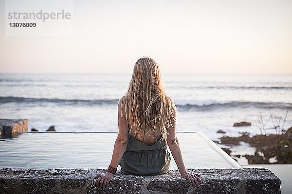 Rückansicht einer Frau  die auf einer Stützmauer an einem künstlichen Teich gegen das Meer und den klaren Himmel sitzt