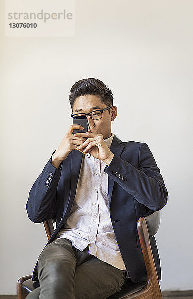 Geschäftsmann benutzt Mobiltelefon  während er im Büro auf einem Stuhl an der Wand sitzt