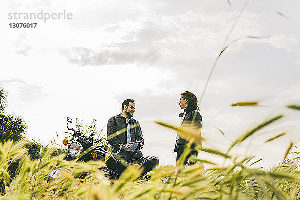 Glückliches Paar unterhält sich  während es mit dem Motorrad auf einem Grasfeld gegen den Himmel steht