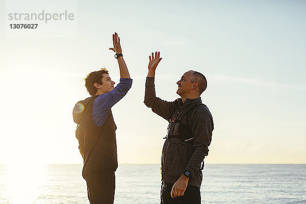 Glückliche Vater und Sohn  die bei Sonnenuntergang ein High-Five gegen Meer und Himmel machen